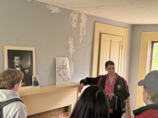 A woman speaks to a group of students, next to a map.