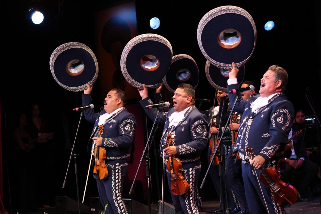 Mariachi performers sing into microphones while holding their hats in the air