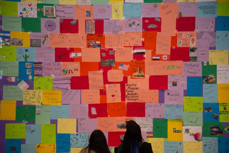 Two students stand in front of a large heart made of post-it notes