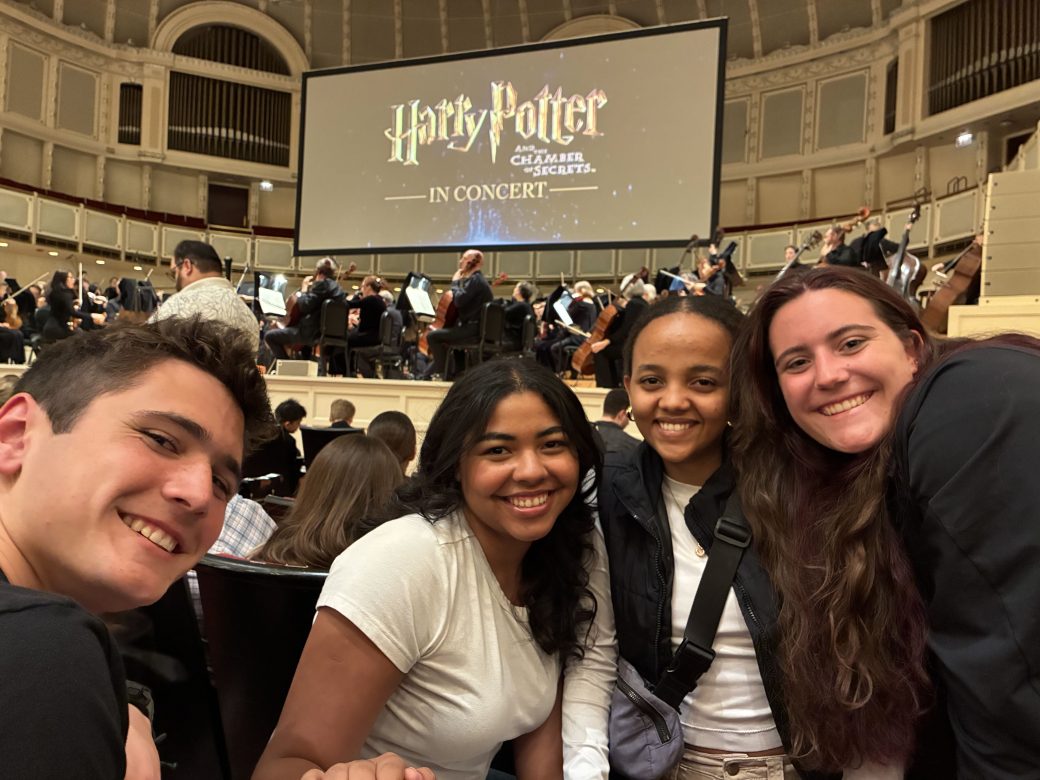 Four students smile in front of a large orchestra and screen that reads "Harry Potter"