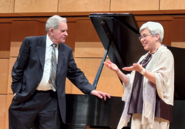 Man in suit and tie and woman talking next to piano