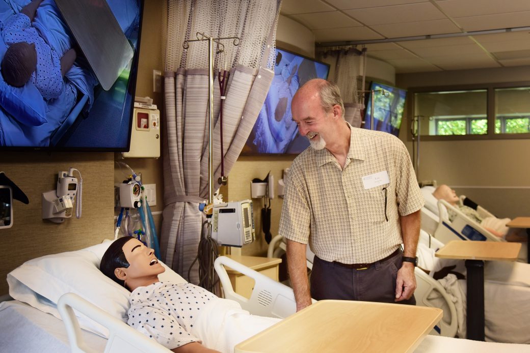 man stands over nursing manikin in hospital bed