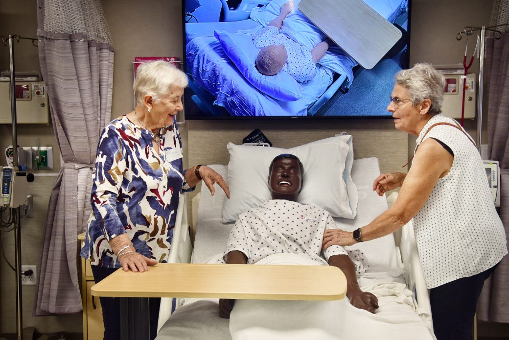 two older nursing alumni consult each other over a new nursing manikin in a hospital bed