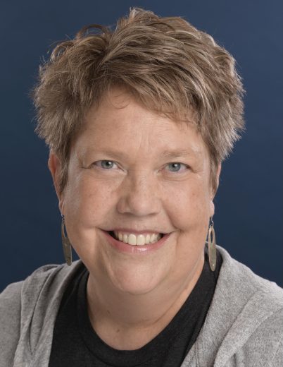professional headshot of Sue Conrad Howes. Conrad Howes in front of a dark blue backdrop wearing a black top and a grey cardigan.