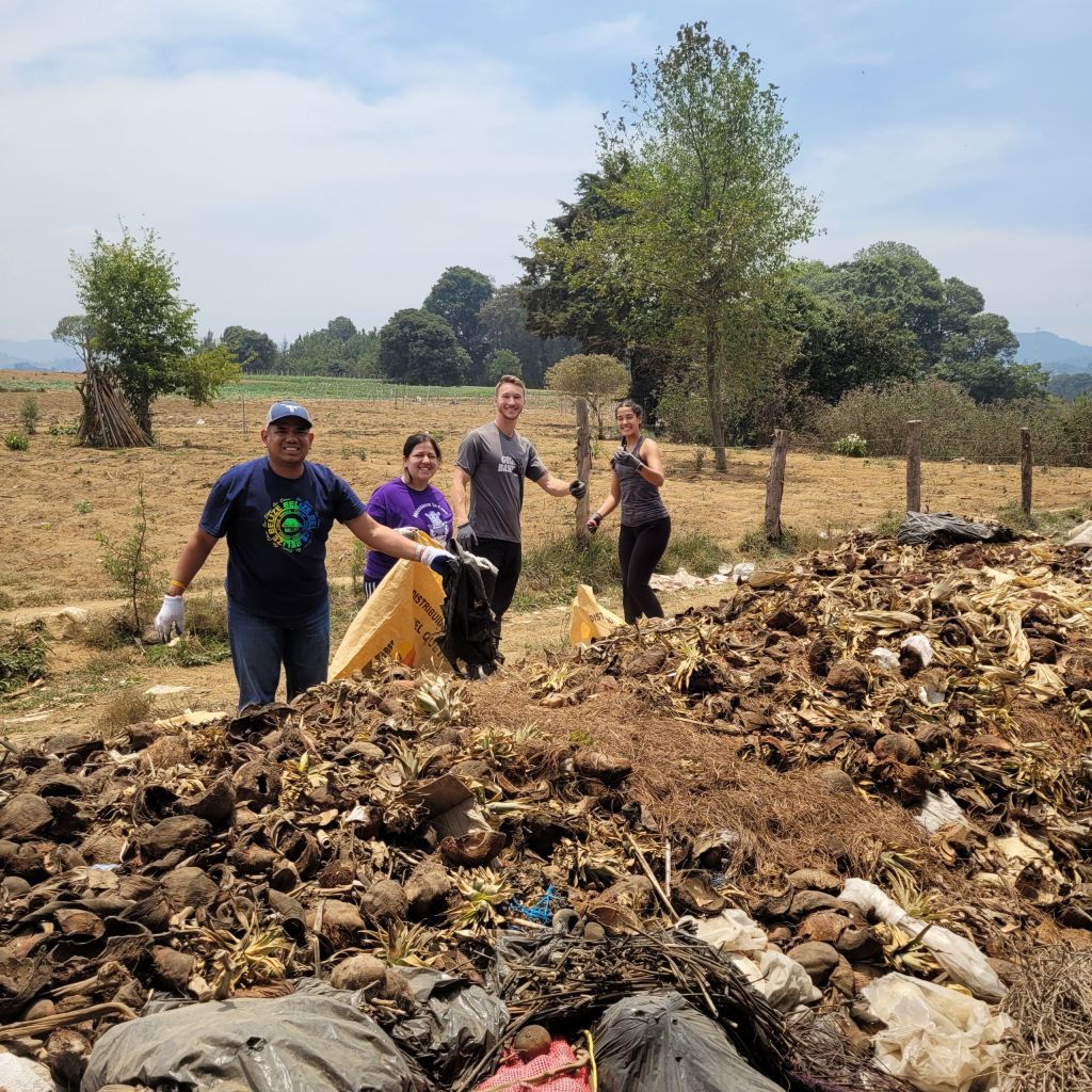 Four individuals smile behind a pile of dirt, plastic, and other waste. They are holding burlap bags and wearing gloves.