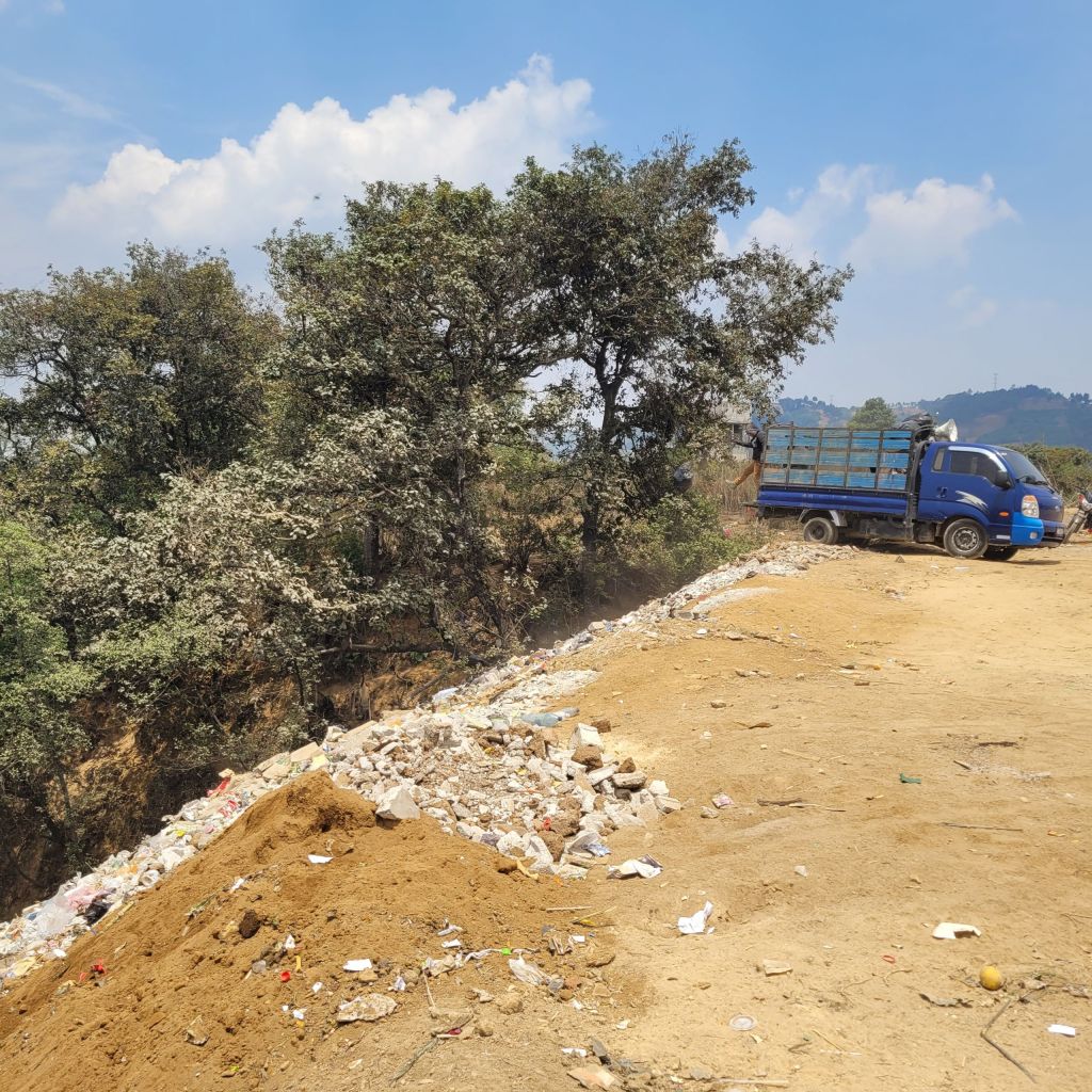 A blue dump truck sits at the edge of a ravine with trash along the edge and trees in the background.