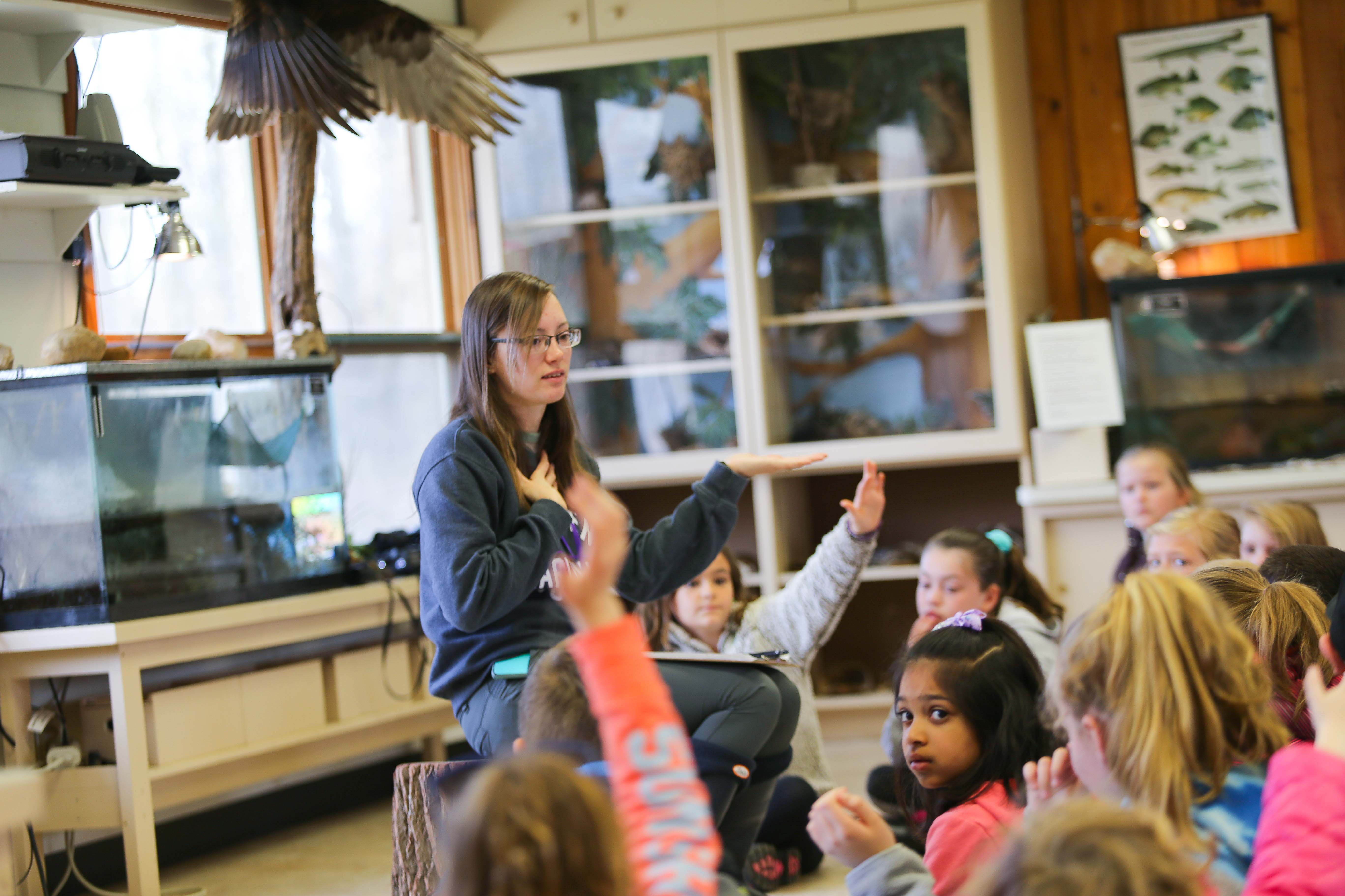 Young children sit on the floor raising their hand facing a woman who appears to be talking with her hands. There are displays and snake tanks in the background.