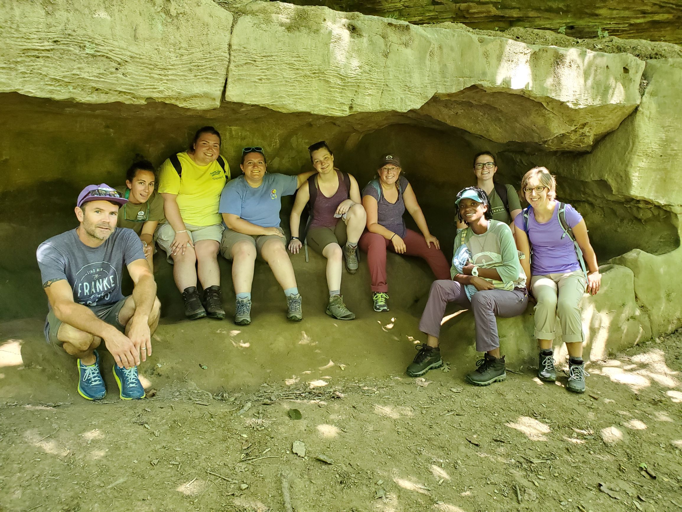 Ten students sit and smile at the camera underneath a large, tan rock that hangs over them.