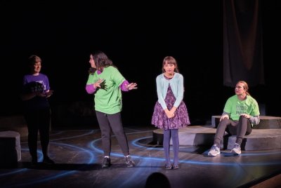 Four student stand at the front of the stage in a line, engaging with each other but facing the audience.