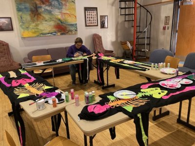 A single person sits at a large square of long tables covered in the painted and drying skeleton costumes.