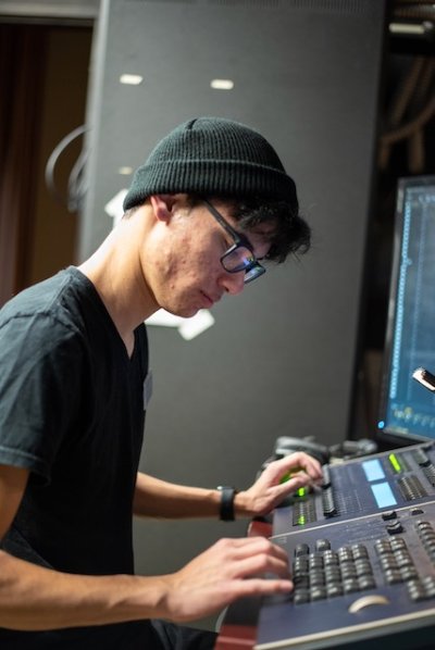 A student wearing dark clothing and a dark colored beanie stands at a switchboard