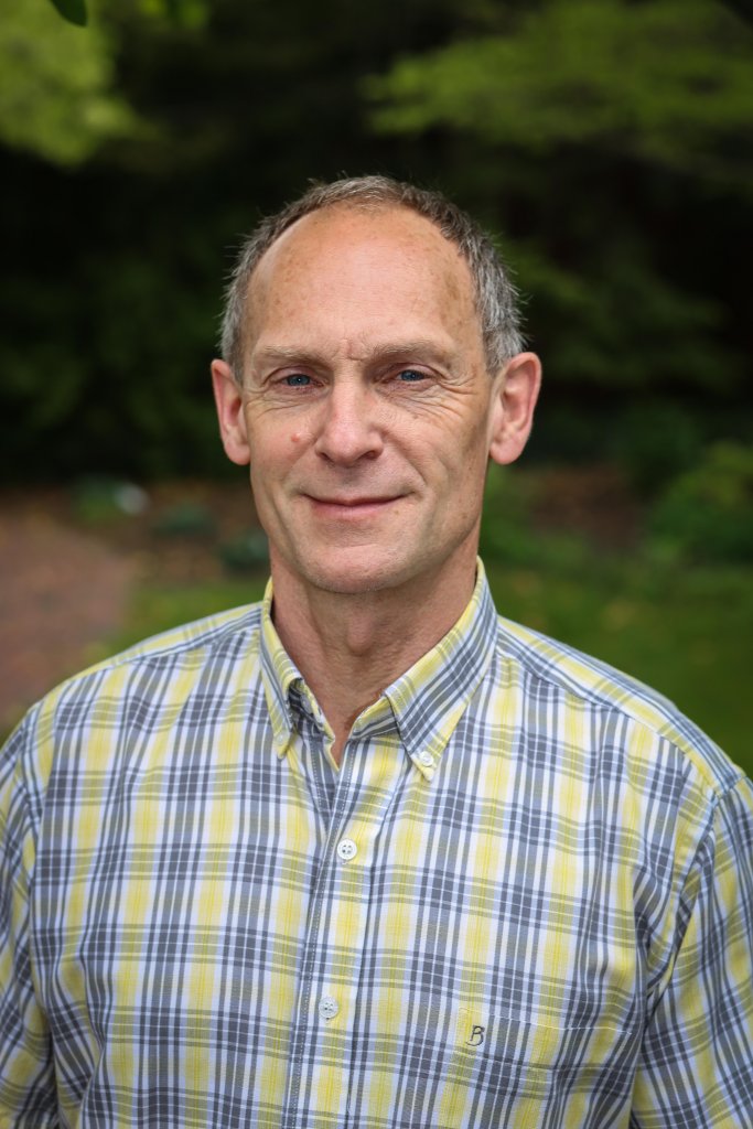 Man in a blue and yellow striped shirt smiling in front of a forest