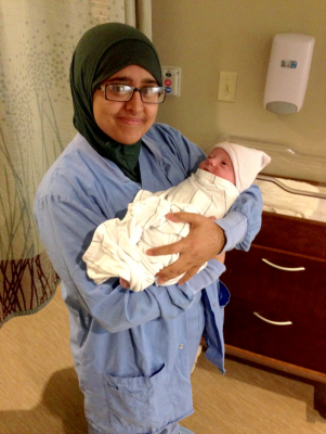 Woman in blue scrubs and a green hijab holding an infant in a white cloth