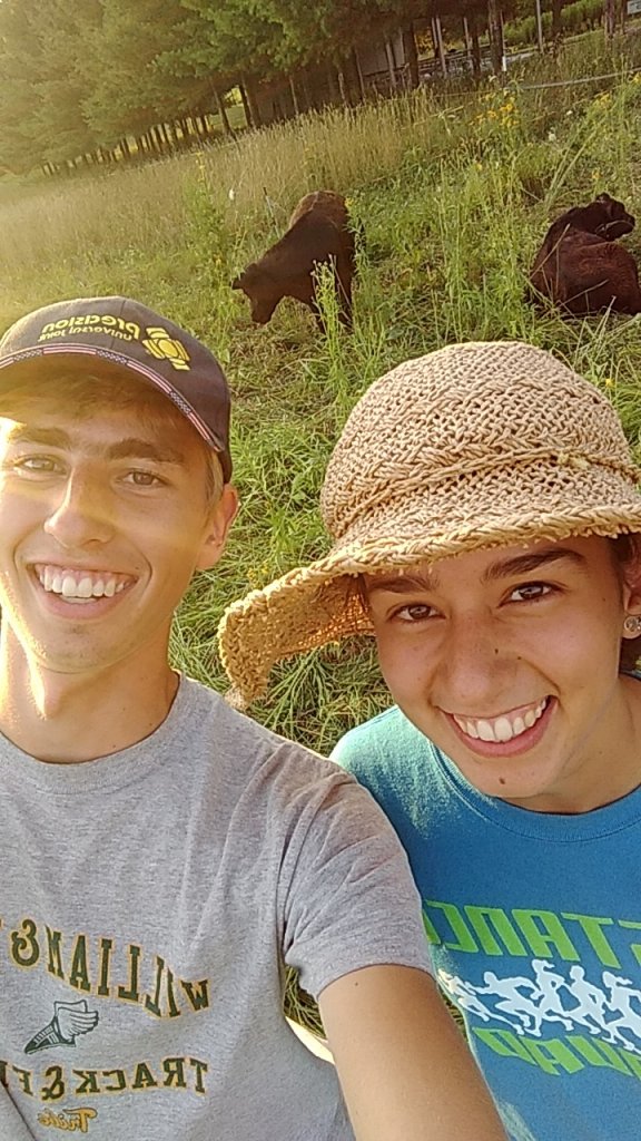 Selfie of two students posing in a field with two black cows behind them