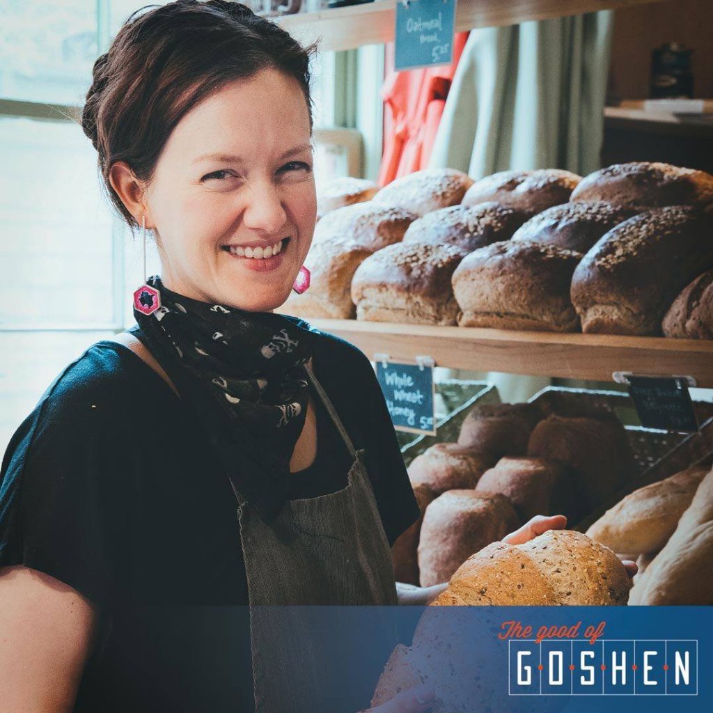 Woman wearing a green apron smiling, holding a loaf of bread