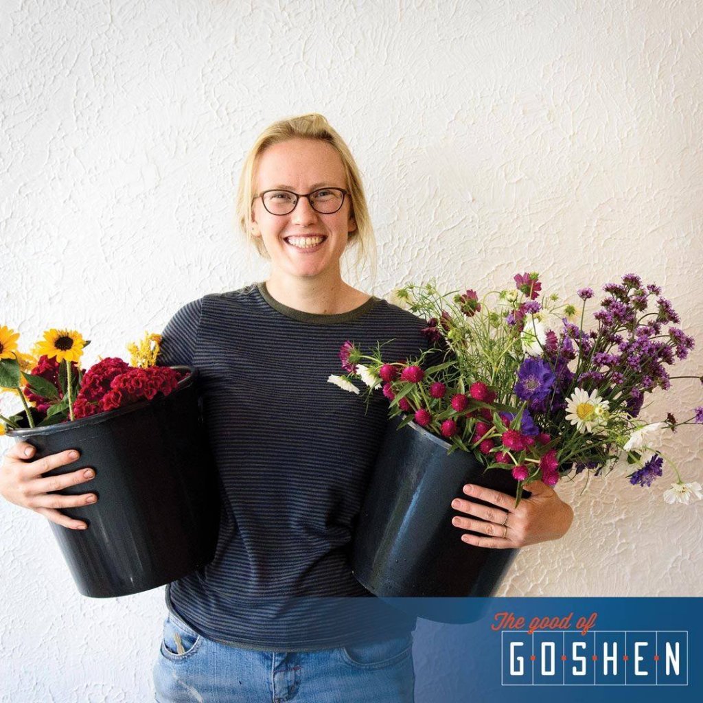 Woman in a black shirt, glasses smiling, holding two black plastic containers full of flowers
