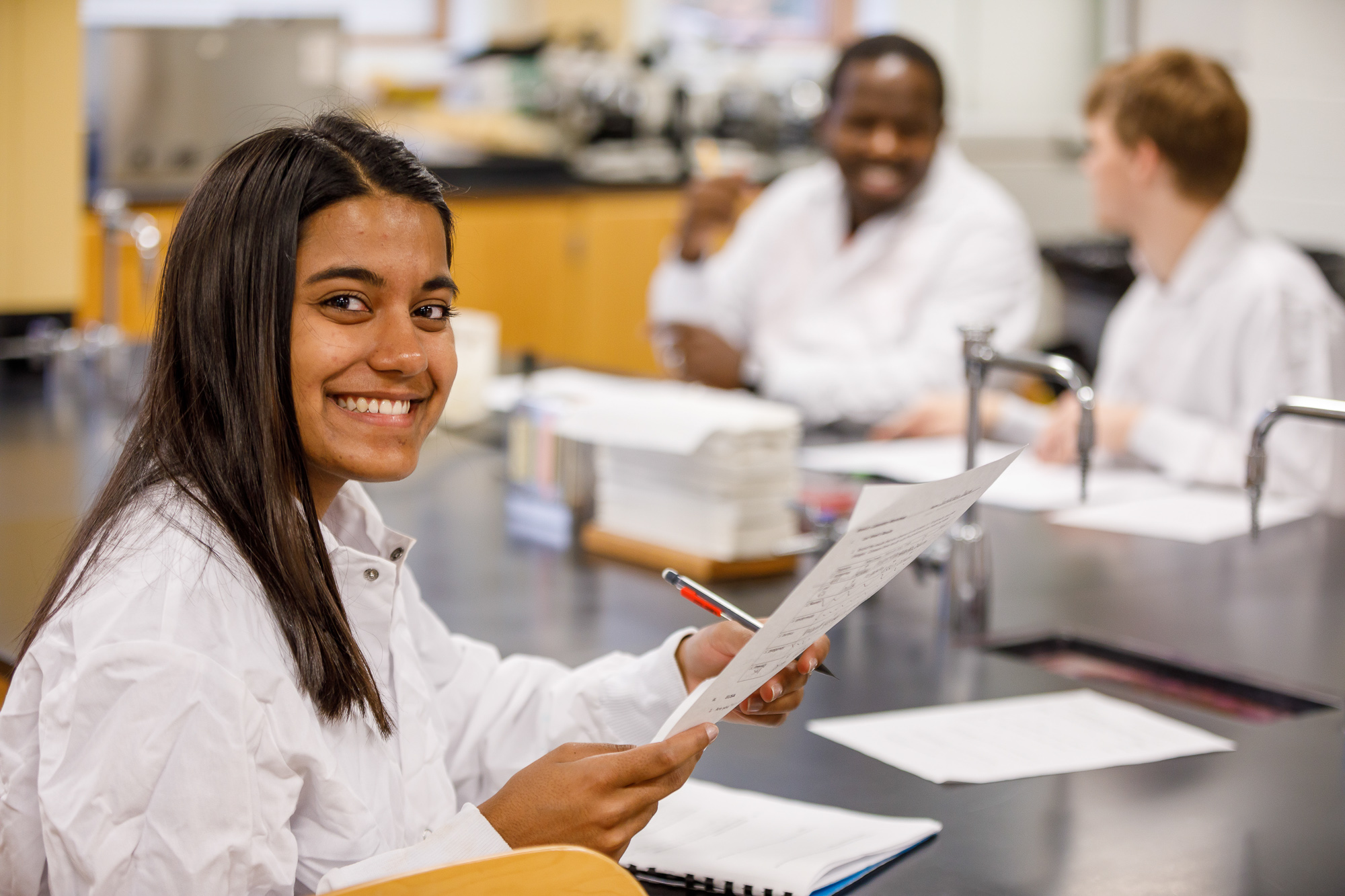 Woman in a lab coat smiling while holding a paper, two students talk behind her