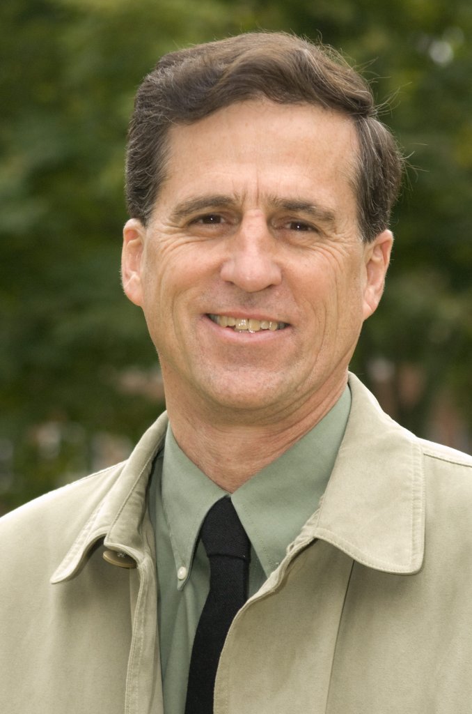 Man in a tan jacket, green button up shirt and black tie smiling