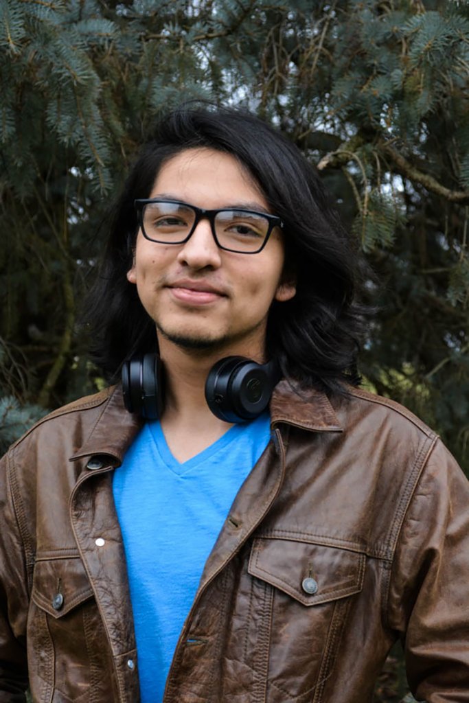 Man wearing a brown leather jacket, blue shirt, glasses and black headphones around his neck smiling