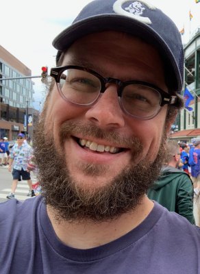 Headshot of a man in a t shirt, baseball cap and glasses smiling