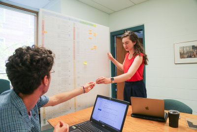 Student in a red shirt handing a sticky note from a white board to a professor sitting at a desk