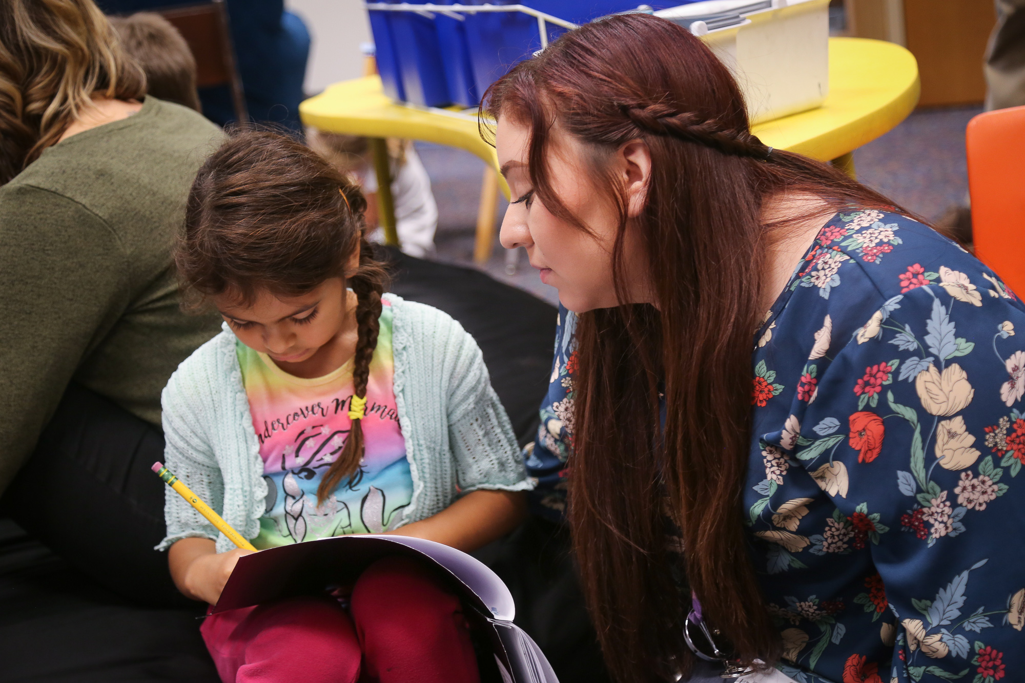 Student in a blue shirt with flower patterns working with a elementary student writing in a notebook