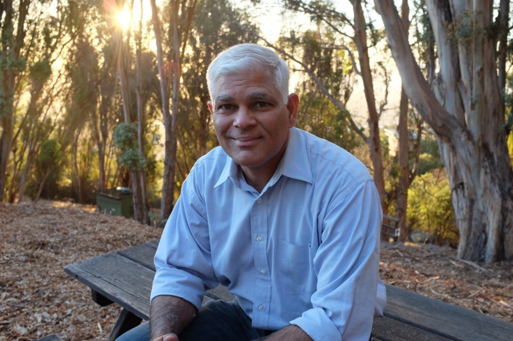 Man wearing a light blue button up shirt sitting on a bench surrounded by trees, smiling