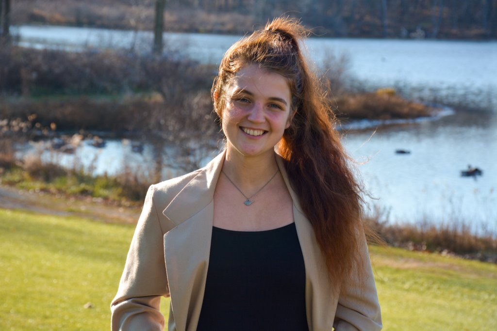 Woman wearing a tan jacket and black shirt smiling in front of a body of water