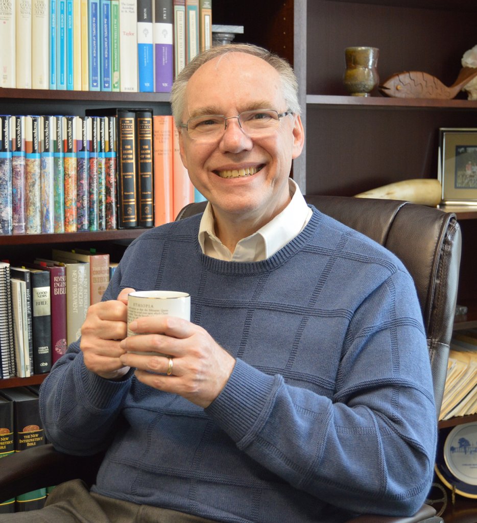 Man wearing a blue sweater, holding a mug sitting in office next to bookshelf smiling