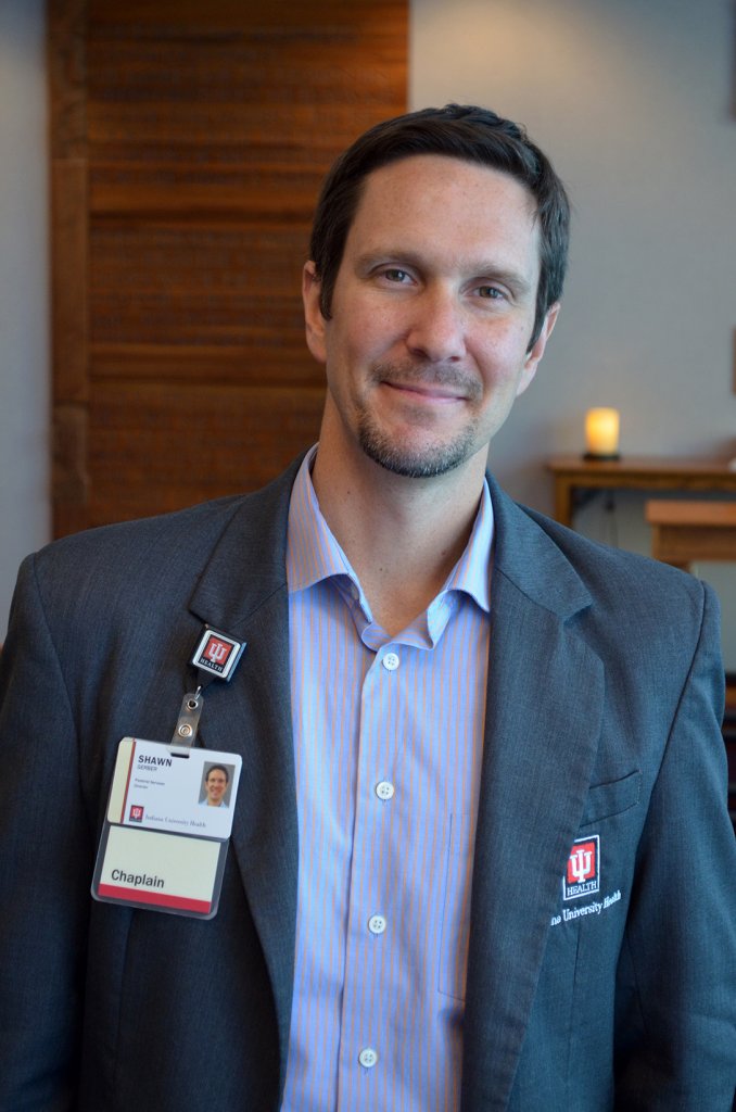 Man wearing a dark blue jacket, white button up shirt and an Indiana University Health ID badge smiling in an office with a candle