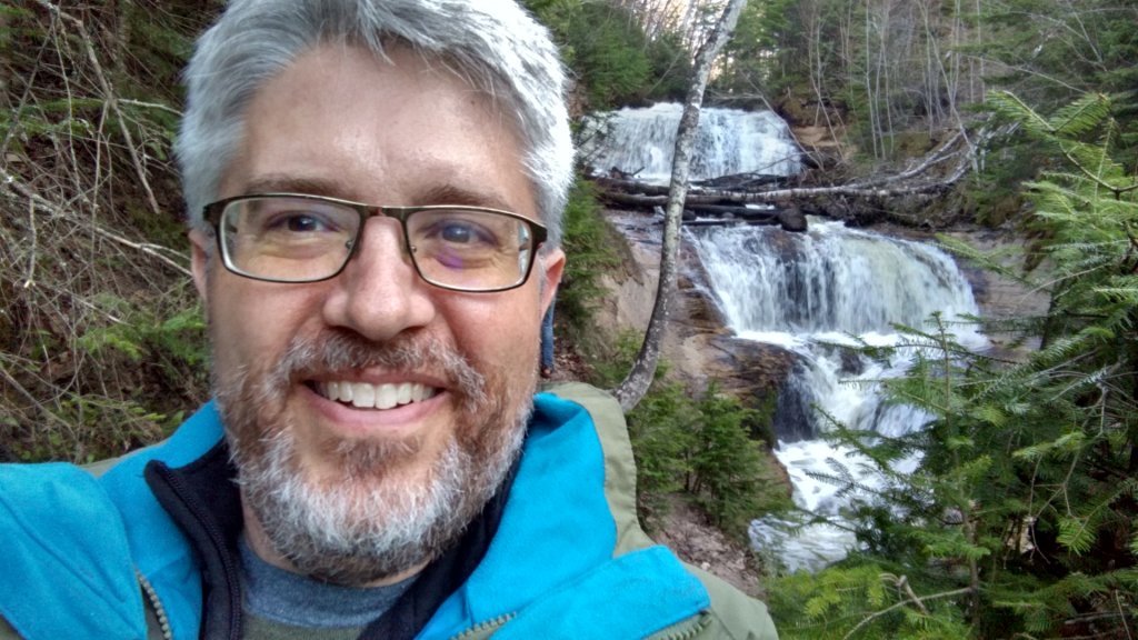 Man wearing a light blue and gray jacket and glasses smiling in front of a small waterfall surrounded by trees.