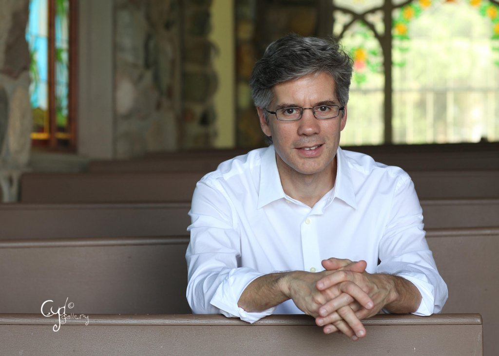 Man wearing a white long sleeve shirt, glasses, sitting in a church pew smiling. In the background is a glass stained window