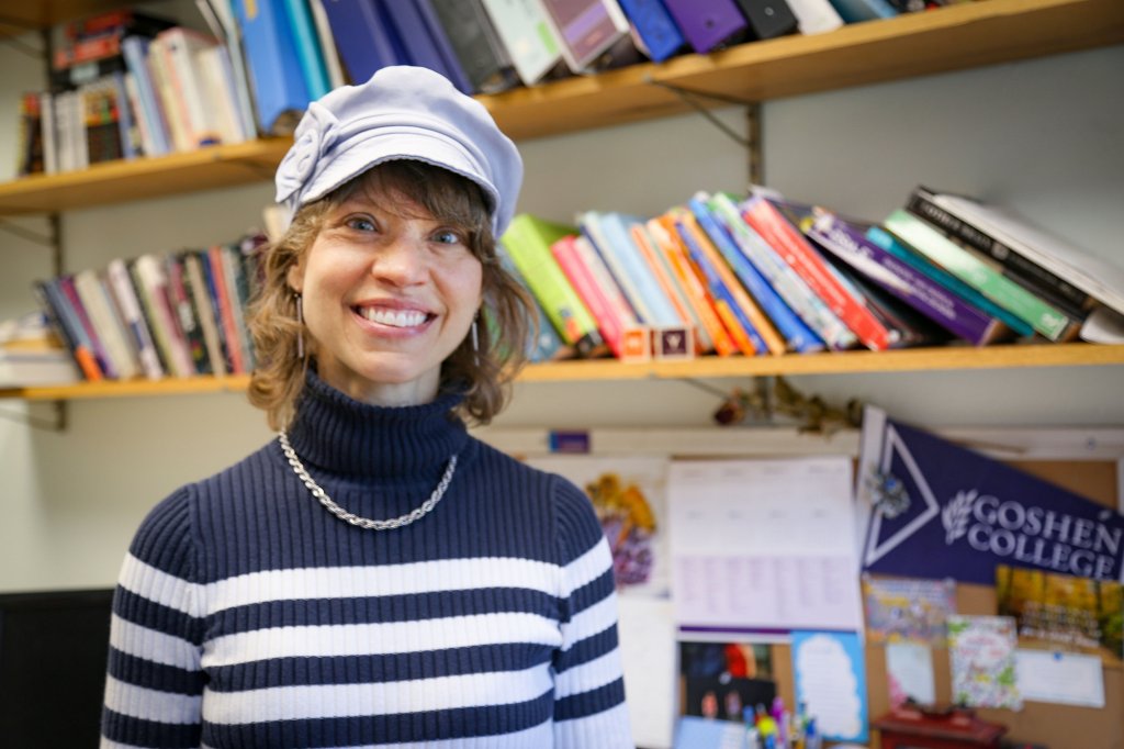 Woman wearing a dark blue and white striped turtleneck, chain necklace and a gray hat smiling in an office