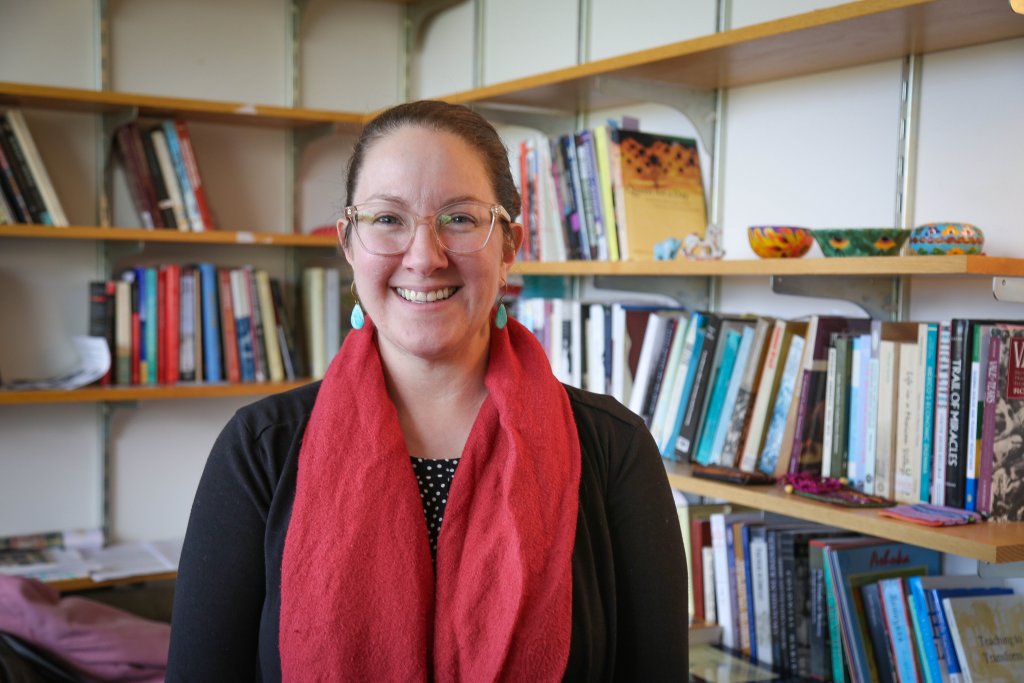 Woman wearing a black shirt, red scarf around her neck, tear shaped turquoise ear rings and glasses smiling in an office