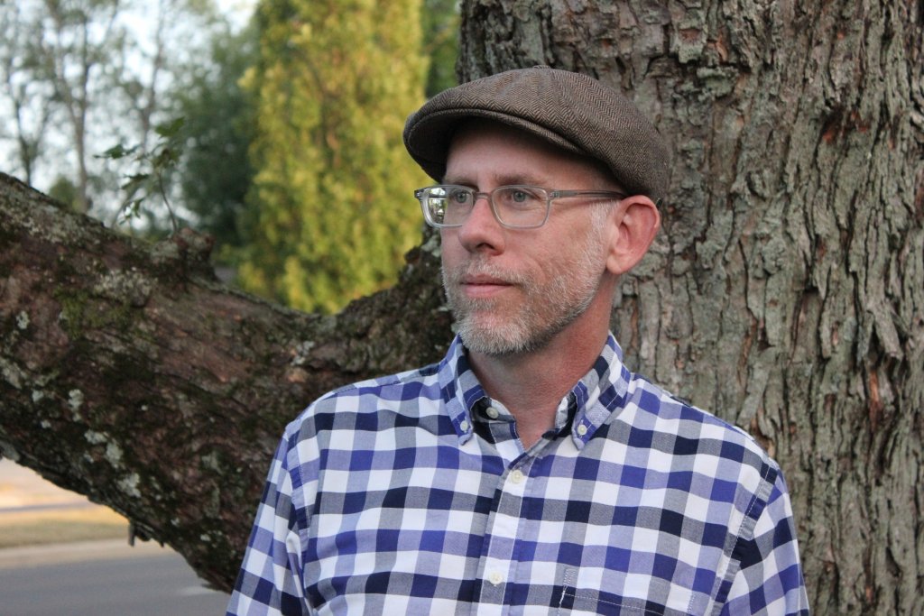 Man in a checker pattern shirt of dark blue, navy blue and gray, glasses and a tan hat standing by a tree.