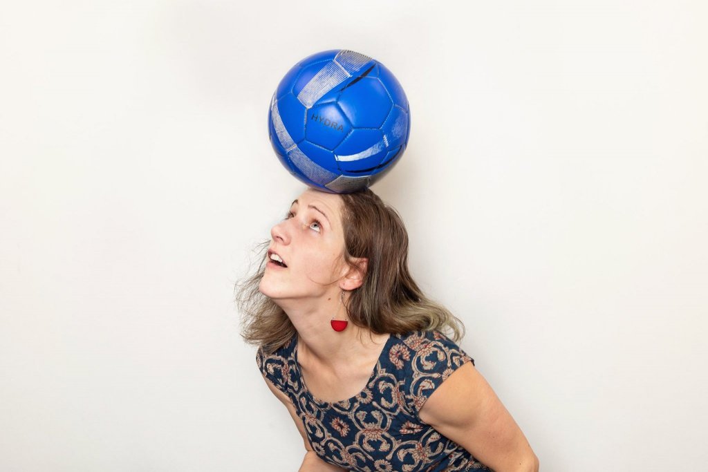 Woman in a dark blue shirt with tan patterns wearing red semicircle ear rings, balancing a dark blue and silver volleyball on her head