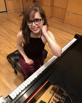 A woman with glasses smiling, sitting by a piano, left elbow resting on the piano.