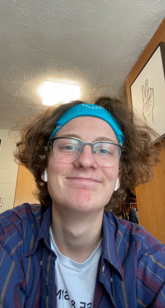 Man wearing a dark purple shirt with red and yellow stripes, light blue headband and glasses smiling in a GC dorm room
