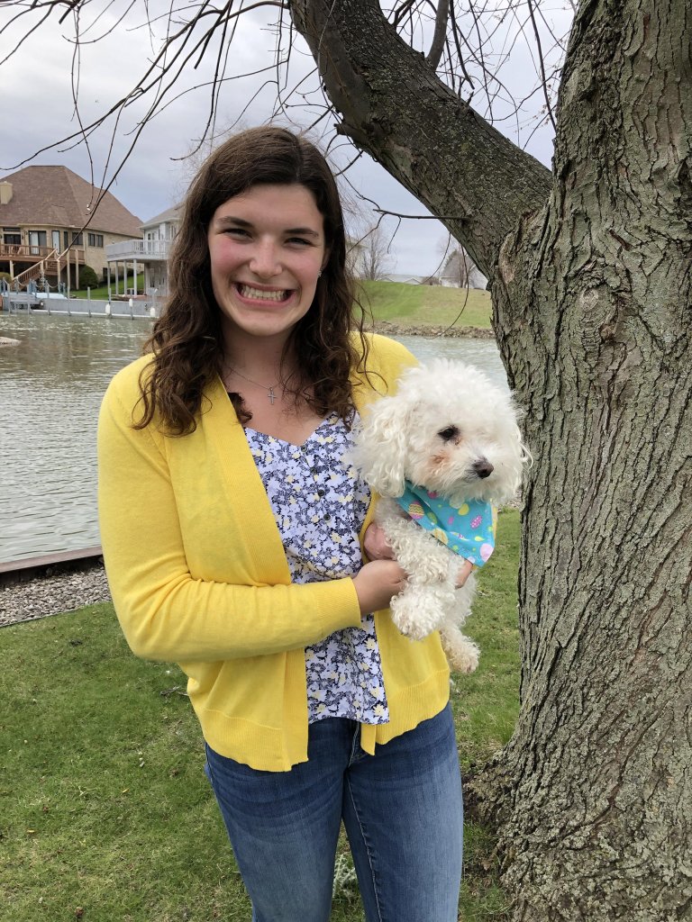 Woman smiling, wearing a yellow blazer and holding a white fluffy dog by a body of water.
