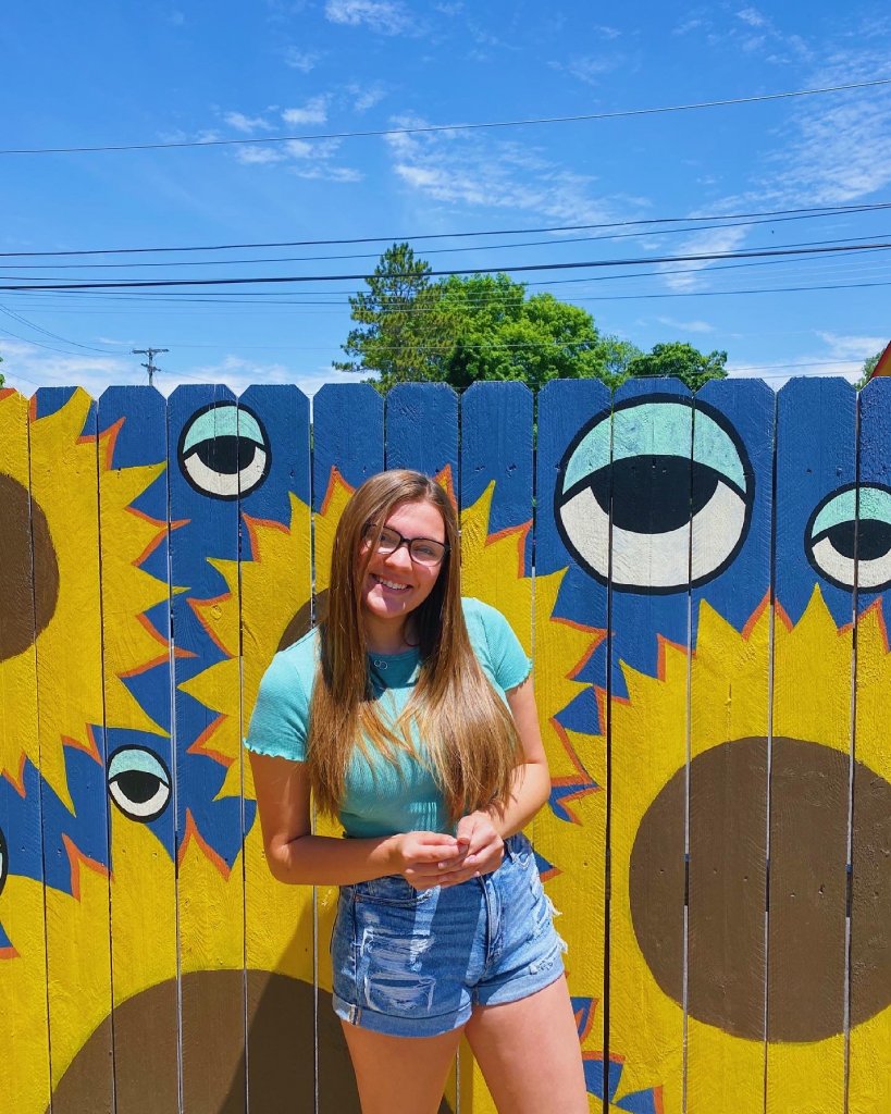 A woman in a light blue t-shirt wearing glasses, smiling, standing in front of a wood fence with giant yellow sunflowers and eyes painted on it.