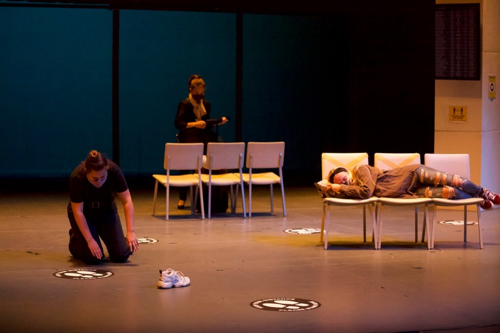 Photo of three actors on stage. One is laying across three white chairs, another in the background looking at another set, and one to the left kneeling by a social distancing sticker.