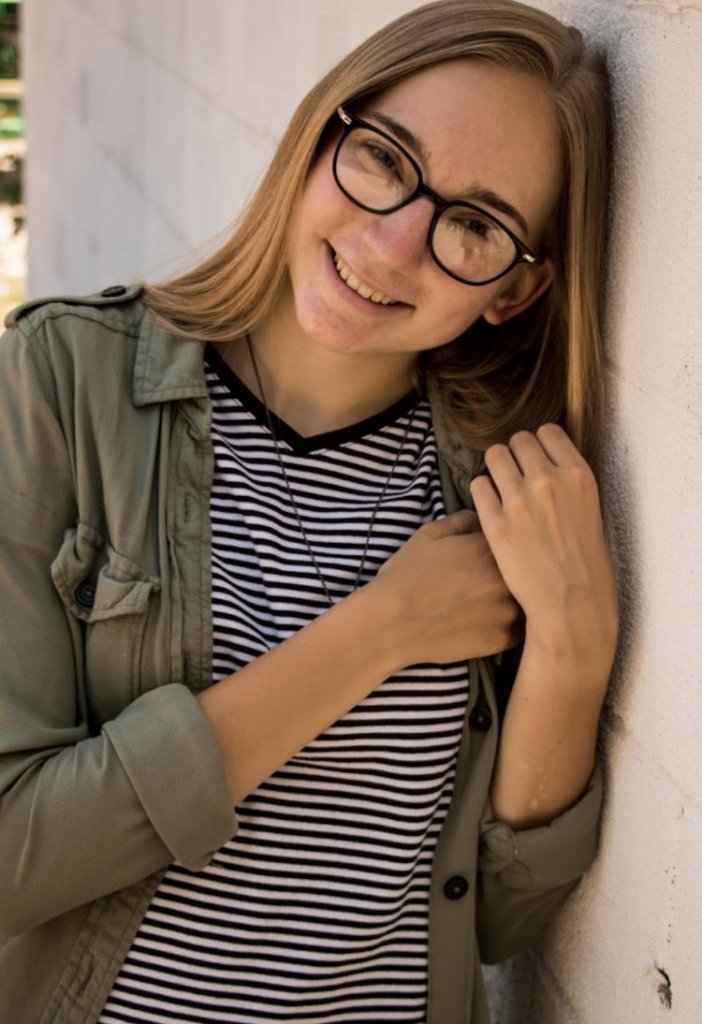 Woman in dark green jacket, glasses smiling, leaning against a white brick wall