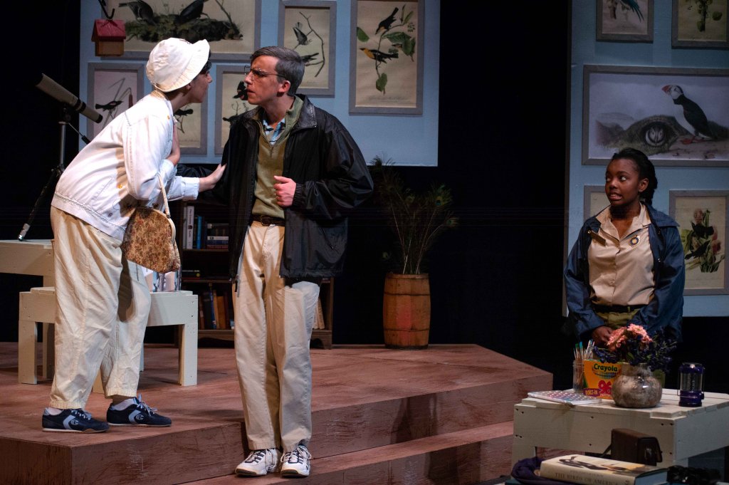 Three students acting; two as older people, talking to each other on the steps that make up a large part of the set, while the other actor watches their exchange.