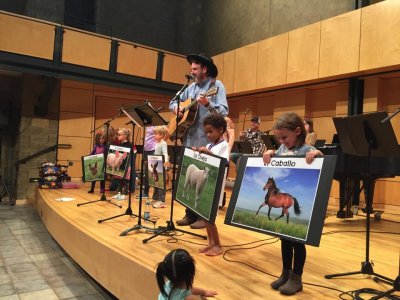 Man singing with a guitar on stage with children who are holding pictures of animals with the animal names in Spanish