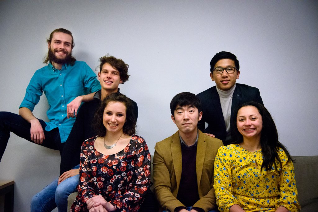 Two females and Four males sitting and leaning on a couch for a group picture