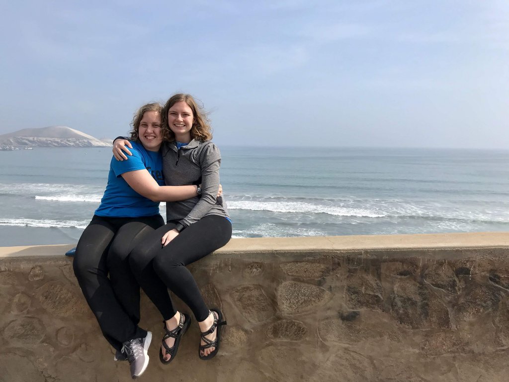 Two sisters sitting on a ledge that over looks the water and a small mountain in the back ground.