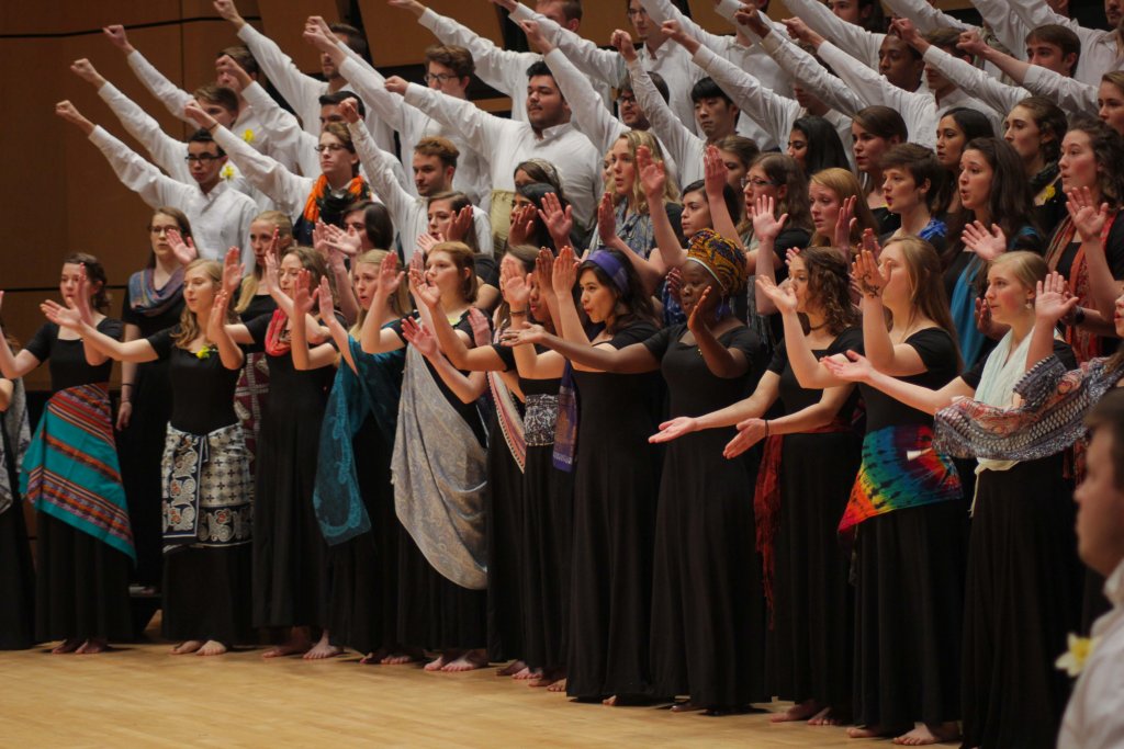 Men's choir, dressed in white shirts and holding their fists in the air, and Women's choir, dressed in black with colorful scarves, singing and clapping