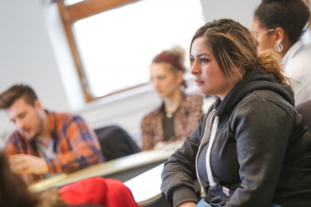 Female student wearing gray sweatshirt in class, with blurred classmates behind her