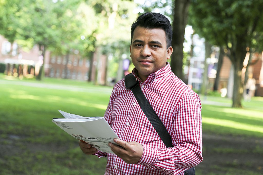 Naun Cerrato in a red and white stripped buttoned up shirt- he is holding a booklet while wearing a shoulder backpack