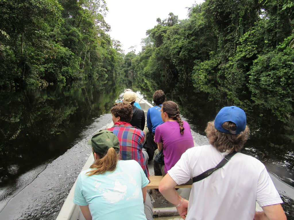 View from behind six students in a canoe on a river, surrounded by green trees